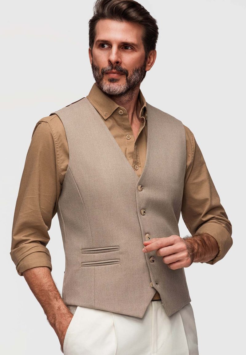 Homme barbu portant un gilet beige sur une chemise marron retroussée, ajustant le bouton du gilet, debout devant un fond neutre.