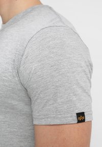 Grå bomulds t-shirt med korte ærmer, der har et let struktureret stof og et lille sort mærke med et logo på venstre ærme.