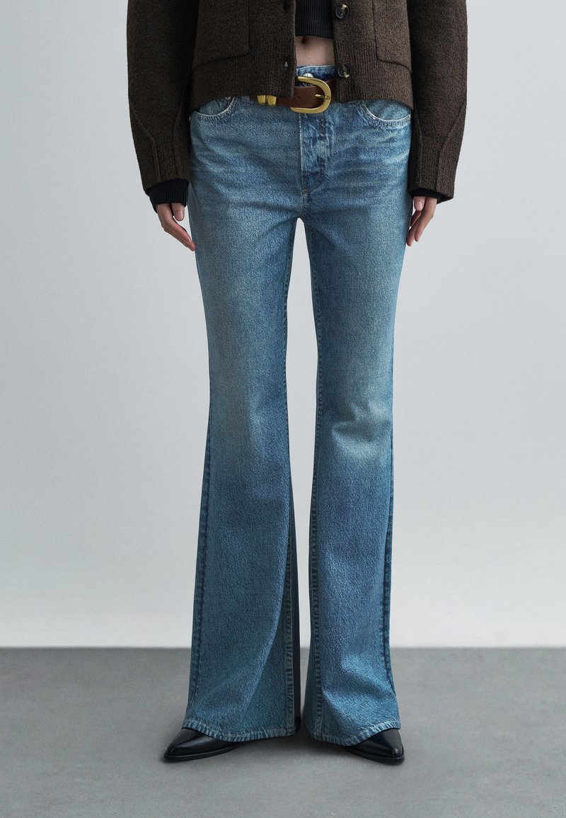 Jeans azuis claros com corte flare e acabamento desgastado, com um design clássico de cinco bolsos e um cinto castanho. O tecido é denim macio.