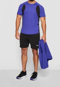 Camiseta deportiva púrpura con acentos laterales negros, pantalones cortos negros y zapatillas negras. Sosteniendo una chaqueta azul, de pie contra un fondo liso.