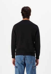 Hombre con cabello castaño lleva una camisa negra de manga larga y jeans cargo azules, de pie de espaldas sobre un fondo blanco.