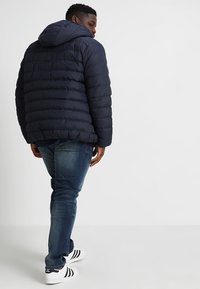 Marineblå oppustelig jakke med hætte, quiltet tekstur og elastiske manchetter, parret med blå jeans og hvide sneakers med sorte striber.