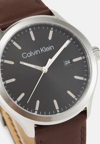 Reloj Calvin Klein con caja redonda de acero inoxidable, diseño de esfera oscura, manecillas plateadas, correa de cuero marrón y display de fecha a las 3 en punto.