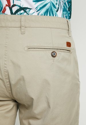 Beige katoenen korte broek met een achterzak voorzien van een bruin leren label en een blauwe knoop. Soepele textuur met subtiele stiksel details.