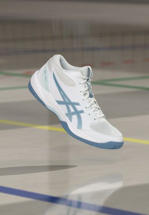 Zapatilla deportiva Asics blanca y azul claro con cordones, suspendida en el aire sobre una pista cubierta con líneas de delimitación de colores.