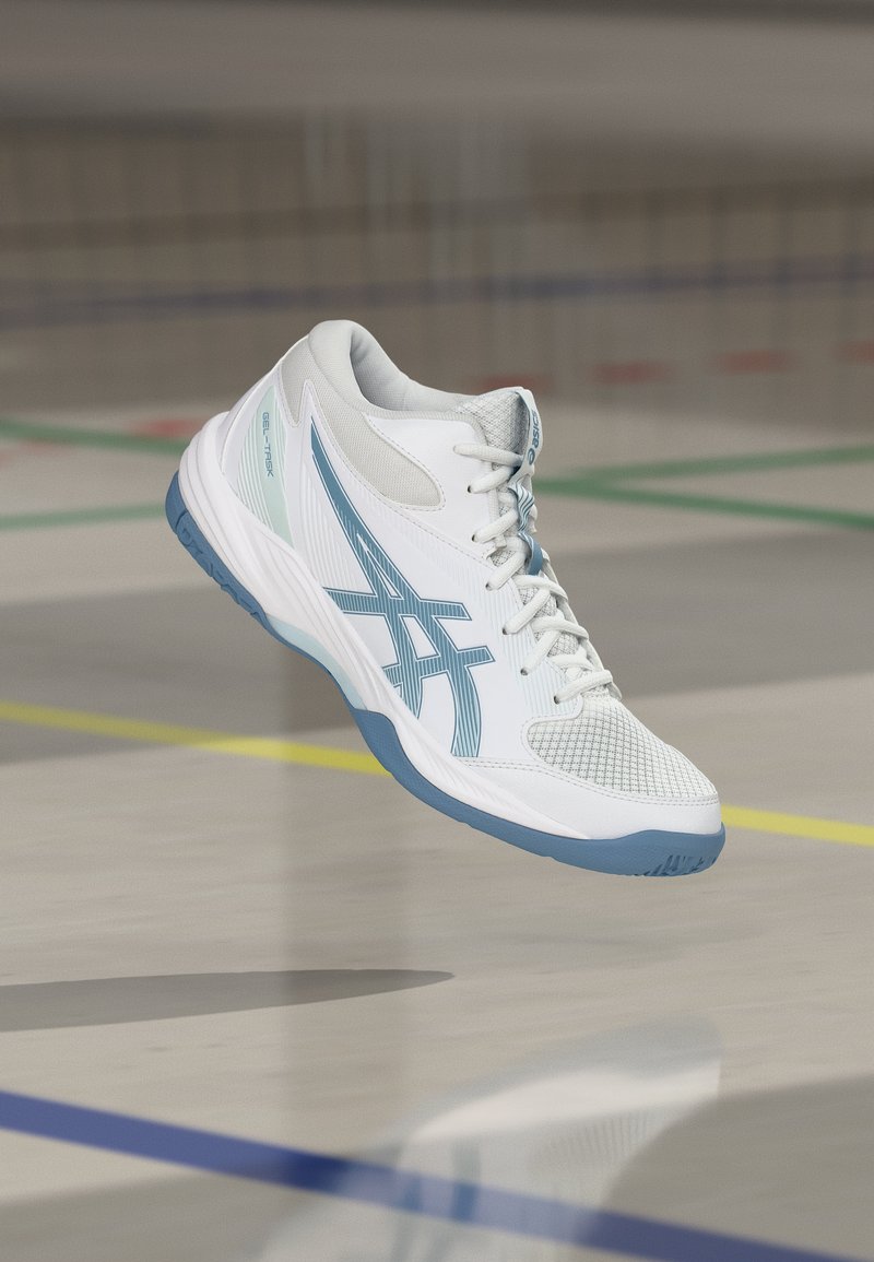 Λευκό και γαλάζιο αθλητικό παπούτσι Asics με κορδόνια, στον αέρα πάνω από εσωτερικό γήπεδο με χρωματιστές γραμμές ορίων.