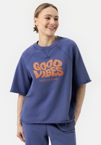 Paarse korte mouwen sweatshirt van textuurstof met een grote oranje print aan de voorkant waarop staat "GOOD VIBES STATE OF MIND".