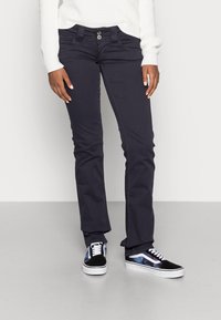 Persoon in zwarte slim-fit broek, witte ribbeltrui en zwart-witte sneakers met een paarse sterrenhemelprint, staande op een grijze vloer.
