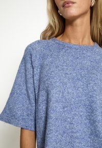 Zils adīts džemperis ar īsām piedurknēm, apaļu kakla izgriezumu un mīkstu, teksturētu apdari. Ar raglanu piedurknēm un delikātu heathered rakstu.