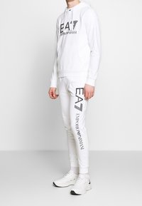 Bílá mikina s kapucí a odpovídající tepláky s černým logem "EA7 Emporio Armani". Hladký bavlněný materiál, přiléhavý design a elastické manžety.