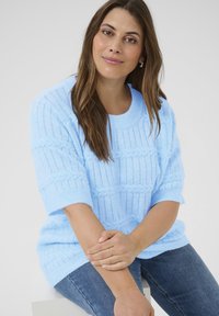 Suéter tejido de color azul claro con un patrón de cables, con mangas cortas y escote redondo. Lleva puesto con jeans azules, con las manos descansando sobre el regazo.