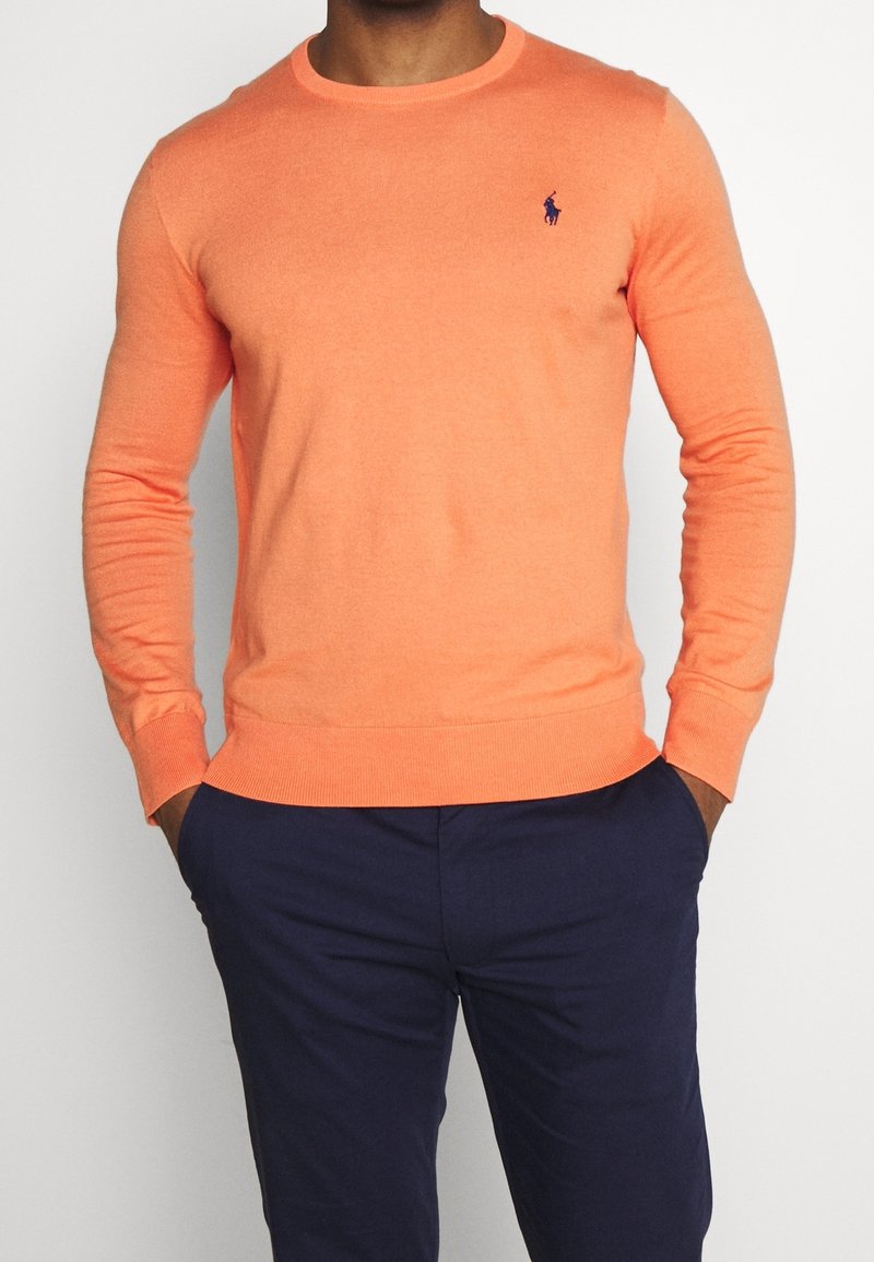 Orange stickad tröja med rund hals och långa ärmar. Har en liten lila logga på bröstet. Bärs över mörknavybyxor.