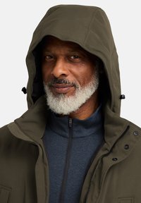 Veste isolante vert olive avec capuche, présentant un avant zippé, un tissu texturé et des fermetures à pression. Portée sur un haut bleu à col montant.