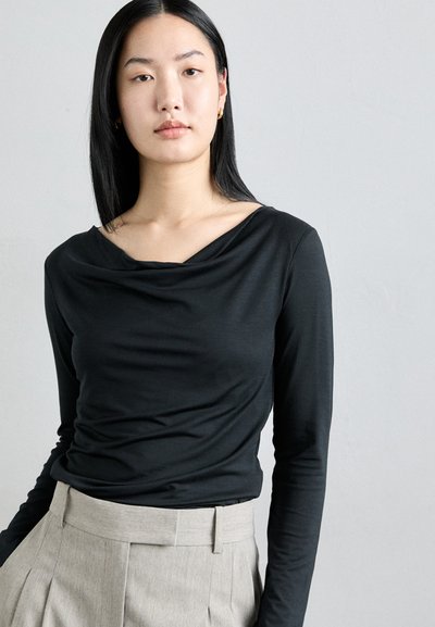 Black long-sleeve top with a draped neckline, smooth texture, and fitted design, paired with light gray high-waisted trousers.