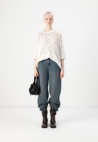 Pull blanc en tricot taille oversize avec un motif géométrique, associé à un jean bleu ample et des bottes marron jusqu'au genou ; portant un sac noir à motif.