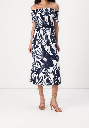 Femme portant une robe midi blanche épaules dénudées avec un imprimé feuilles tropicales bleu marine et des sandales à talons fines marron clair.