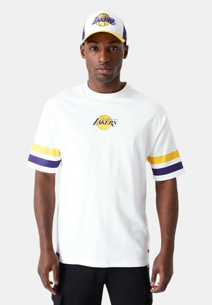 Hombre con camiseta blanca de Los Angeles Lakers con rayas amarillas y moradas en las mangas y gorra a juego de los Lakers, de pie frente a un fondo gris.