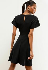 Donna con coda di cavallo che indossa un vestito nero a maniche corte con dettaglio a chiave sulla schiena, orecchini a cerchio dorati e un braccialetto.