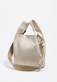 Beżowa skórzana torba o miękkim fasonie, z teksturowanym wykończeniem, szerokim materiałowym paskiem i metalicznym elementem na zapięciu.