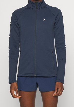 Personne portant une veste zippée bleu marine avec un col montant et du texte sur les manches, associée à un short bleu marine assorti.