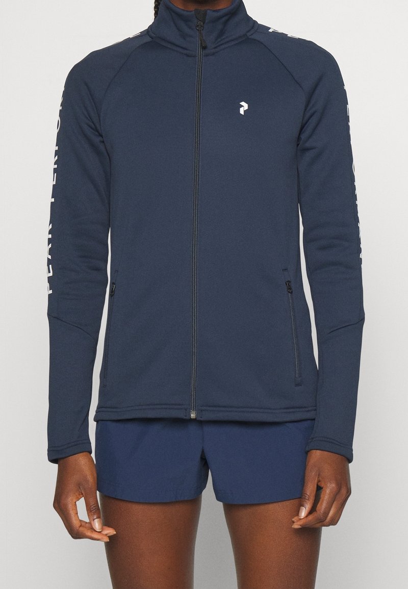 Personne portant une veste zippée bleu marine avec un col montant et du texte sur les manches, associée à un short bleu marine assorti.
