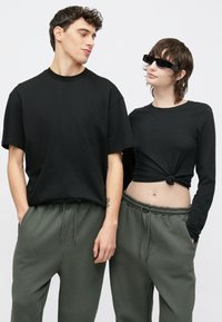 Fekete pamut pólók, bő szabásúak; a bal oldali modellen rövid ujjú póló, a jobb oldalin hosszú ujjú, deréknál megkötve. Zöld szabadidőnadrágok.