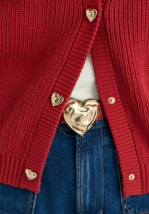 Czerwony prążkowany kardigan z złotymi guzikami w kształcie serc oraz klamrą w pasku, noszony na białej bluzce i niebieskich dżinsach. Tkanina o fakturze.
