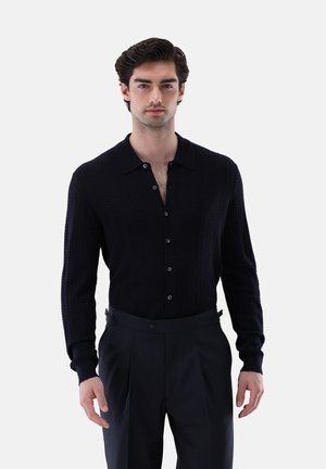 Mann mit dunklem, welligem Haar trägt ein schwarzes, strukturiertes, geknöpftes Langarmhemd und schwarze, maßgeschneiderte Hose und steht vor einem schlichten Hintergrund.