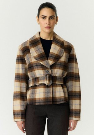 Femme portant une veste ceinturée à carreaux marron et beige sur un top noir et un pantalon foncé, debout devant un fond clair uni.