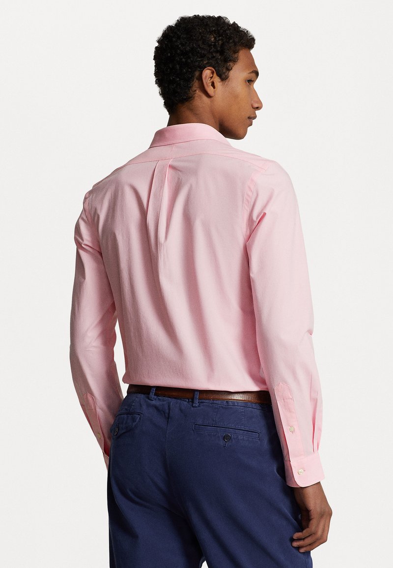 Camicia button-up rosa chiaro con piega sul retro, maniche lunghe e polsini con bottoni, abbinata a pantaloni blu navy e una cintura marrone.