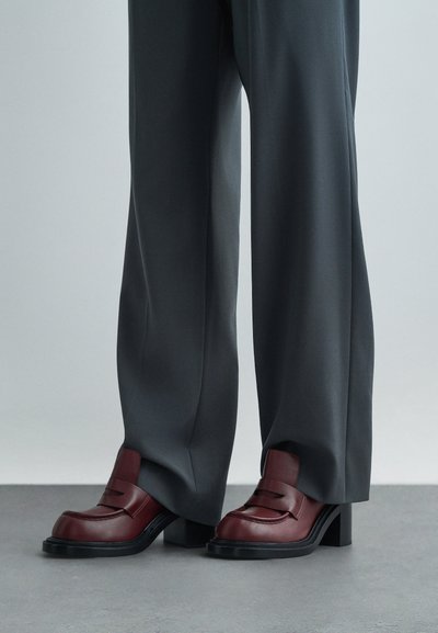 Burgundské kožené mokasíny s nízkým blokovým podpatkem a švy, spárované se šedými kalhotami zakrývajícími vršky bot.