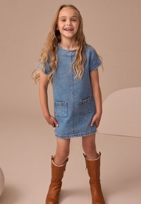Šviesiai mėlynos spalvos džinsinė suknelė trumpomis rankovėmis ir dviem priekinėmis kišenėmis, papildyta rudais aukštakulniais batais iki kelių. Lygi tekstūra ir kasdienė stiliaus dizainas.
