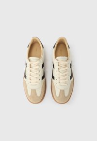 Hellbeige Sneaker mit schwarzen Streifen und braunen Wildledereinsätzen. Abgerundete Spitze, flache Schnürsenkel und Gummisohle. Minimalistisches Design und klassische Form.