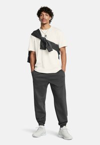 Conjunto casual masculino: camiseta crema con logotipo, pantalones de chándal gris oscuro y un suéter gris atado alrededor de los hombros, combinado con zapatillas blancas.