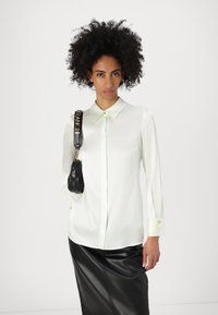 Blusa verde chiaro a maniche lunghe con colletto classico, chiusura a bottone e polsini. Abbinata a una borsa a tracolla nera imbottita e una gonna di pelle.