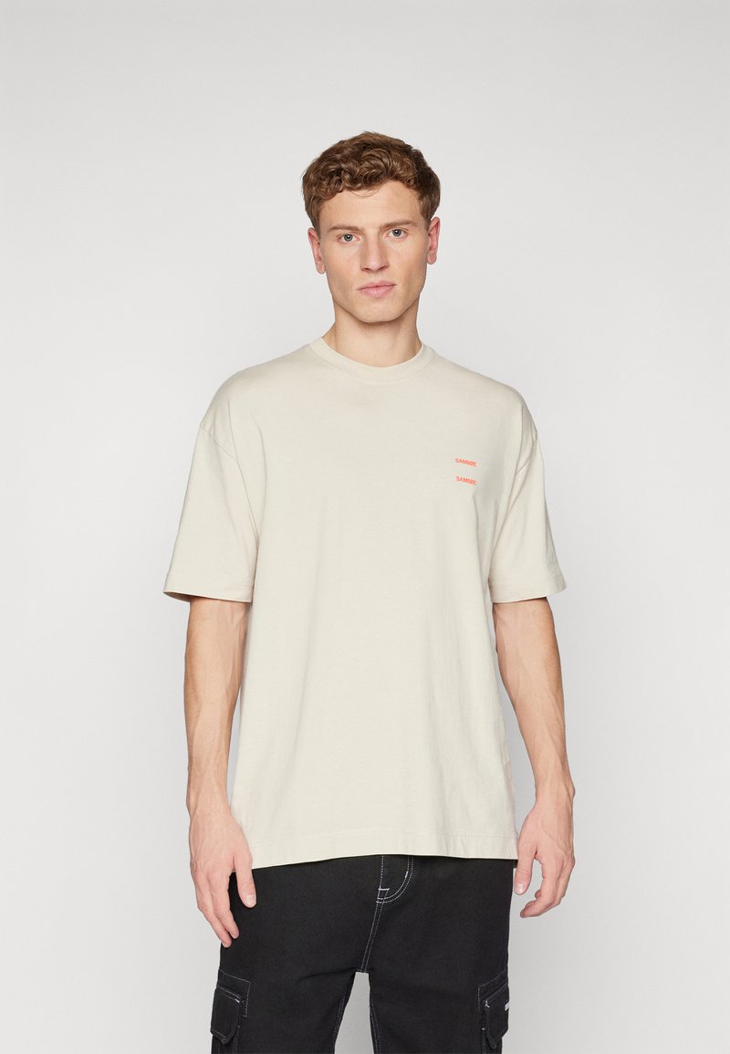 T-shirt en coton beige avec un col rond et des manches courtes, présentant deux petites impressions rouges "SANDRO" à l'avant. Coupe décontractée classique.