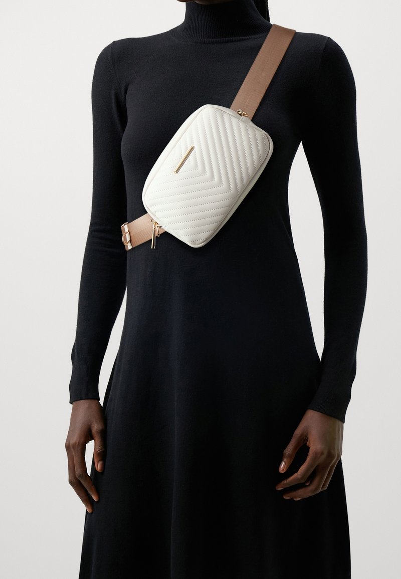 Prošívaná bílá syntetická crossbody taška obdélníkového tvaru s béžovým řemínkem, zdobená zlatými kovovými prvky a diagonálními vzory prošití.