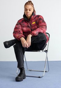 Doudoune à motifs rouge et noir, avec fermeture éclair à l'avant et logo. Associée à un jean noir et des bottes compensées, assise sur une chaise en chrome.