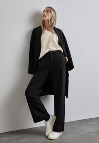 Schwarz weit geschnittene Hose, cremefarbene Rippstrickpullover, langer schwarzer Mantel. Modell trägt weiße Sneakers. Glatte Texturen und klare Linien sichtbar.