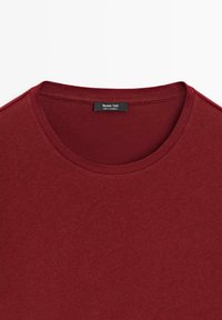 T-shirt bordeaux à col rond avec des coutures apparentes et une étiquette noire cousue à l'intérieur du col indiquant "Massimo Dutti Made in Cambodia".