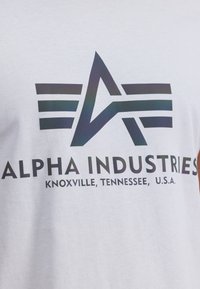 Camiseta blanca con un logotipo negro que contiene una "A" y tres franjas horizontales, el texto dice "ALPHA INDUSTRIES, KNOXVILLE, TENNESSEE, U.S.A."