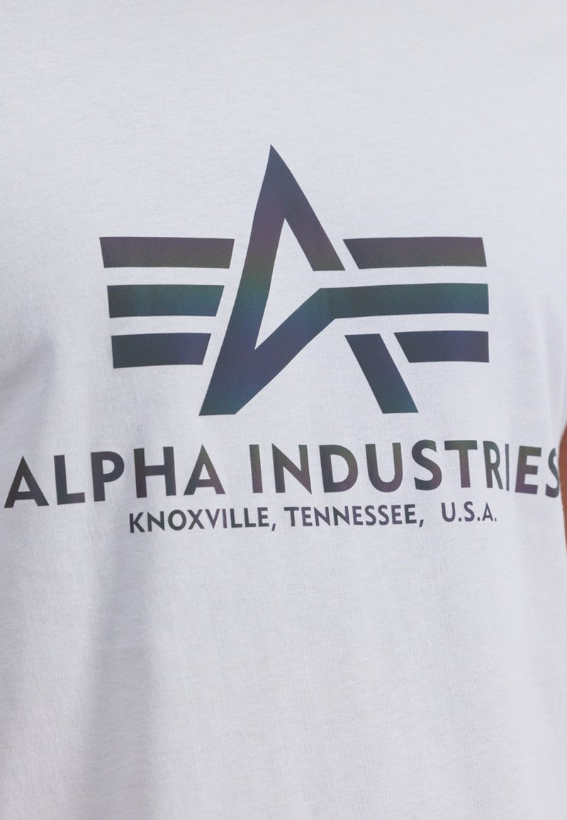 Camiseta blanca con un logotipo negro que contiene una "A" y tres franjas horizontales, el texto dice "ALPHA INDUSTRIES, KNOXVILLE, TENNESSEE, U.S.A."