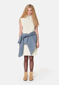 Cremefarbenes ärmelloses Kleid mit geripptem Kragen und drei Knöpfen, kombiniert mit einem blauen gestrickten Wickel um die Taille und tanfarbenen, mit Pelz gefütterten Ankle Boots.