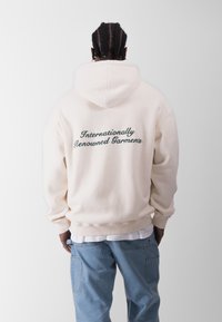 Sudadera color crema con un gran texto en verde cursiva en la parte posterior que dice "Prendas de Renombre Internacional". Cuenta con capucha con cordones y puños acanalados.