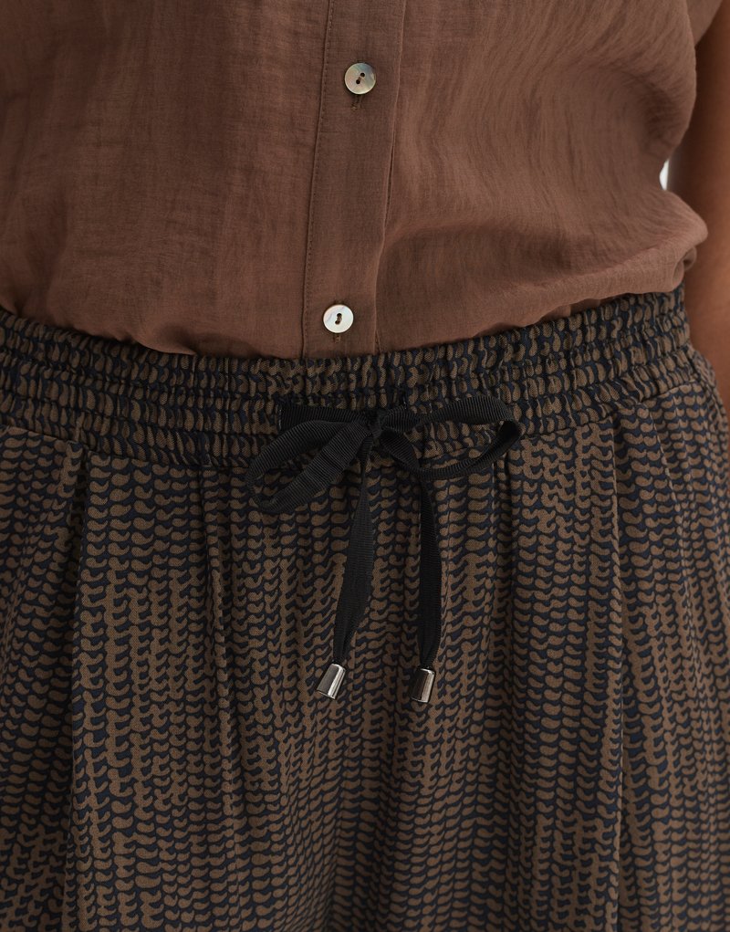 Braune Bluse mit Knopfverschluss über einem schwarz-braun gemusterten Rock mit gerafftem elastischem Bund und Kordelzug. Metallspitzen an den Enden des Kordelzugs.