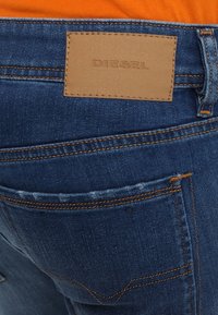 Jeans de denim con un parche de cuero marrón etiquetado como "DIESEL", con costuras naranjas y un detalle de bolsillo trasero desgastado.