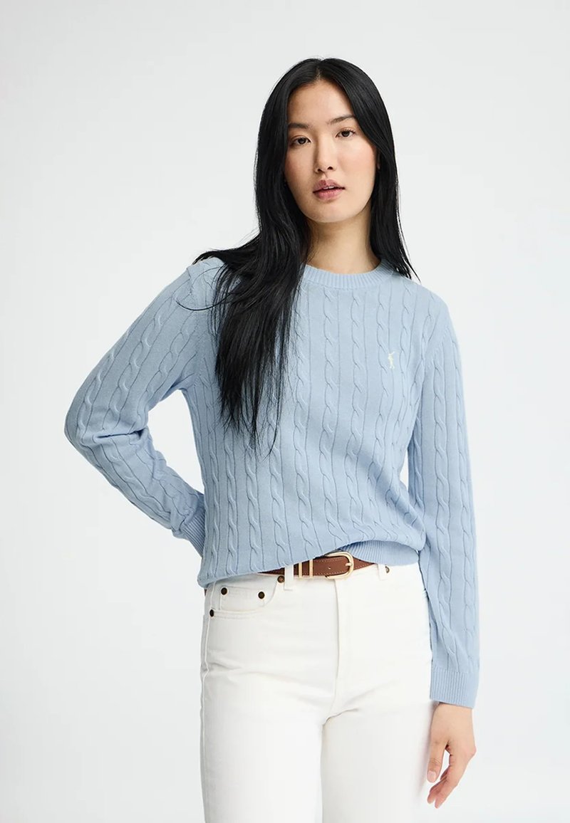 Kvinde med langt sort hår iført en lyseblå kabelstrikket trøje og hvide bukser med et brunt bælte, stående foran en ensfarvet baggrund.