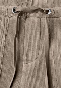 Närbild av beige corduroybyxor med texturerade vertikala ränder och en platt dragrem knuten genom metallöglor i midjan.