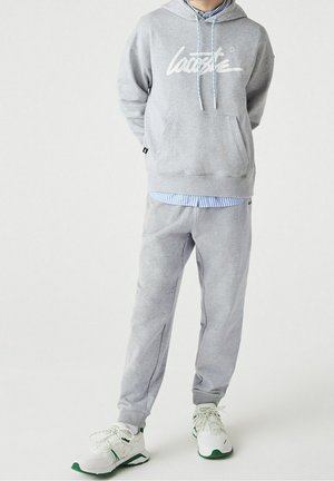 Persoon die een grijze Lacoste hoodie en bijpassende joggingbroek draagt met witte sneakers, staand tegen een effen witte achtergrond.