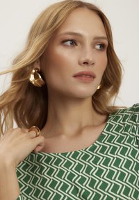 Zielona sukienka z geometrycznymi białymi wzorami, okrągły dekolt, gładka tekstura. Złote kolczyki w kształcie kół i pierścionek dopełniają stylizację. Kolor skóry wydaje się naturalny.